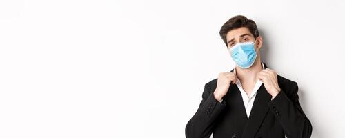Concept of covid-19, business and social distancing. Image of confident handsome man in trendy suit and medical mask, looking sassy, standing against white background photo