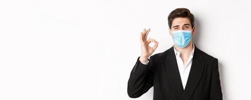 Concept of covid-19, business and social distancing. Close-up of good-looking businessman in trendy suit and medical mask, showing okay sign and winking, standing against white background photo