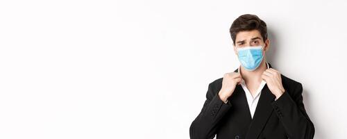 Concept of covid-19, business and social distancing. Image of confident handsome man in trendy suit and medical mask, looking sassy, standing against white background photo