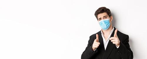 Concept of covid-19, business and social distancing. Close-up of sassy man in trendy suit and medical mask, pointing fingers at camera, standing against white background photo