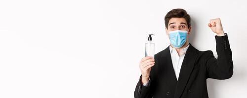 Concept of covid-19, business and social distancing. Close-up of happy man in trendy suit and medical mask, cheering and raising hand up, showing hand sanitizer, white background photo