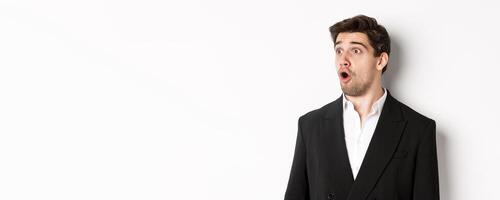 Close-up of impressed guy in trendy suit, open mouth and looking left wondered, standing against white background photo