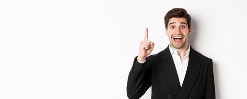 Close-up of handsome businessman in black suit, smiling amazed, showing number one, standing over white background photo