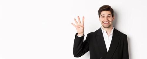 Close-up of handsome businessman in black suit, smiling amazed, showing number four, standing over white background photo