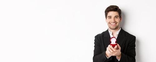 Image of handsome man looking romantic, open small box with engagement ring, making a proposal and smiling, standing against white background photo