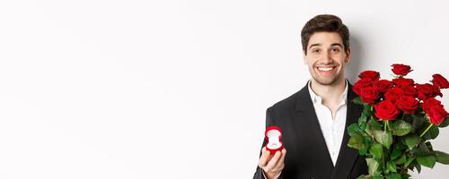 Close-up of attractive man in suit, holding bouquet of roses and engagement ring, making proposal, standing against white background photo