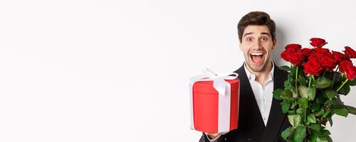 Close-up of handsome bearded man in suit, holding present and bouquet of red roses, smiling at camera, standing against white background photo