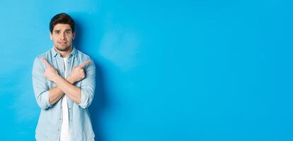 Portrait of nervous guy pointing fingers sideways, looking indecisive and asking for help with choice, showing left and right promo offers, standing against blue background photo