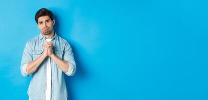 Young guy begging for help, pouting and looking sad while asking for favour, need something, standing against blue background photo