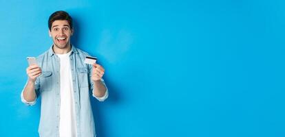Amazed handsome man shopping online, holding mobile phone and credit card, smiling while paying for internet purchase, standing over blue background photo