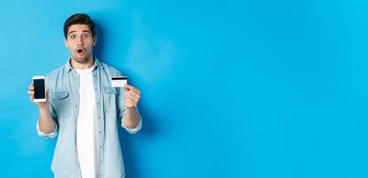 Amazed young man showing mobile cell phone screen and credit card, shop online, standing against blue background photo