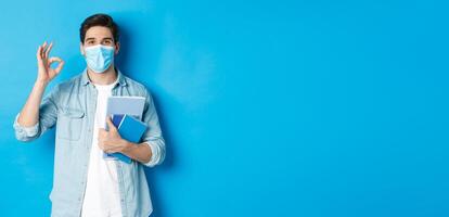 Education, covid-19 and social distancing. Guy student in medical mask looking happy, holding notebooks, showing ok sign, standing over blue background photo