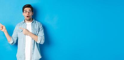Surprised man in glasses pointing fingers left at copy space, asking question about product, looking intrigued, standing over blue background photo