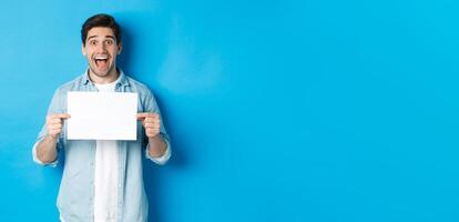 Excited man making fantastic announcement, showing logo or sign on blank piece of paper, standing over blue background photo