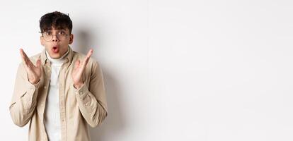 Portrait of surprised hipster guy in glasses checking out awesome promo, say wow and gasping amazed, raising hands up with disbelief, standing on white background photo