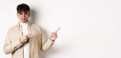 Angry young man frowning and pointing at upper right corner banner, complaining and looking disappointed, standing on white background photo