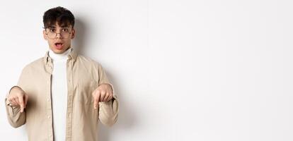 Shocked attractive young man showing something awesome, drop jaw and gasping in awe, pointing fingers down, standing on white background photo