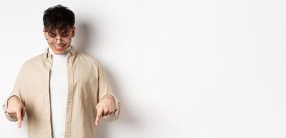 Stylish modern guy in glasses pointing, looking down with happy smile, showing banner, standing on white background photo