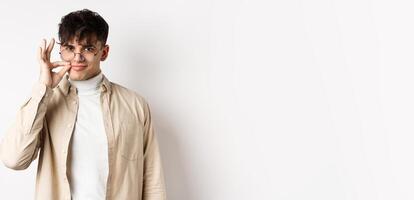 Smiling cunning guy in glasses shut his mouth on zipper, showing seal gesture near lips and smiling devious, keeping a secret, standing on white background photo