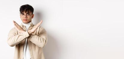 Image of serious young guy in glasses say no, showing cross gesture, make stop sign to disagree or prohibit something, standing on white background photo