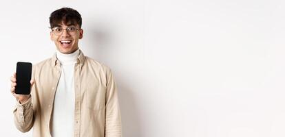 Technology and online shopping concept. Cheerful handsome guy in glasses showing empty smartphone screen and smiling, showing application, standing on white background photo