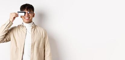 Cheerful young guy smiling real and showing plastic credit card, standing happy on white background, wearing glasses photo