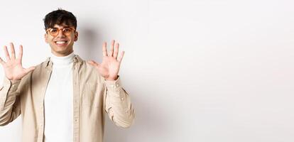 Modern guy in glasses and stylish outfit, showing ten fingers number and smiling, standing on white background photo