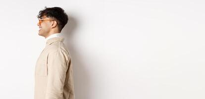 Profile of handsome caucasian man in eyewear, looking at empty space left with cheerful smile, standing on white background photo