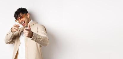 Handsome funny man in glasses smiling, pointing fingers at camera, choosing or inviting you, standing on white background photo