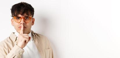 Close-up of grumpy young guy in glasses frowning and shushing, make taboo gesture with finger shuting lips, tell to be quiet, standing angry on white background photo