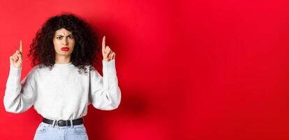 Grumpy young woman with curly hair frowning and grimacing unsatisfied, pointing fingers up at something bad, complaining on company, red background photo