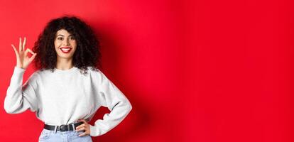 Confident smiling woman in casual clothes, showing OK sign to approve or like product, guarantee good quality, making a compliment, red background photo
