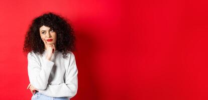 Grumpy young woman with curly hair, looking annoyed or bored at empty space, standing pensive and sad on red background photo