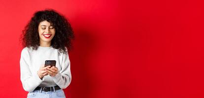 Happy positive girl chatting on phone, reading message and smiling, using social media app, standing on red background photo