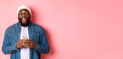 Technology and online shopping concept. Surprised young Black man using mobile phone, looking at camera amazed and happy after reading message, pink background photo