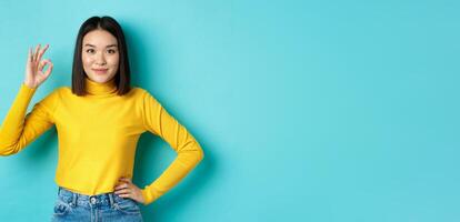 Beautiful smiling asian woman recommend product, showing Ok sign and looking satisfied, standing over blue background photo