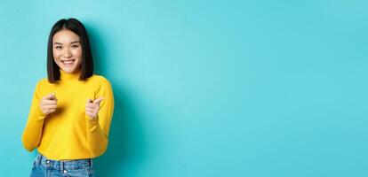 Smiling young asian woman pointing fingers at camera, choosing you, inviting to event, standing happy against blue background photo