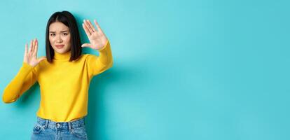 Image of woman feeling uncomfortable in front of camera, cover face with hands and frowning, begging to stop, standing against blue background photo
