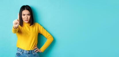 Confident and serious woman tell no, showing extended finger to stop and prohibit something bad, frowning and looking at camera self-assured, standing over blue background photo