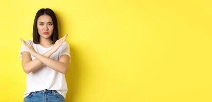 Serious and assertive korean woman showing cross stop gesture, frowning and telling no, prohibit action, disapprove something bad, standing over yellow background photo