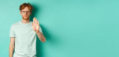 Angry and serious young man with red hair, wearing glasses, showing stop gesture, telling no, disapprove and prohibit something bad, standing over turquoise background photo