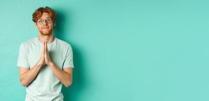 Cute redhead man asking for help, saying please with namaste gesture, pleading or begging for favour, looking silly at camera, standing over mint background photo