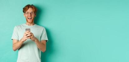 Surprised redhead man in glasses looking amazed at camera after reading promo offer on smartphone, standing against turquoise background photo