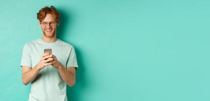 Handsome young man in glasses with red messy hair reading message on mobile phone, smiling and looking at screen, standing over mint background photo