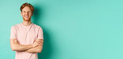 Portrait of friendly-looking young man with red hair and beard smiling and starng satisfied, holding hands crossed on chest, standing over turquoise background photo