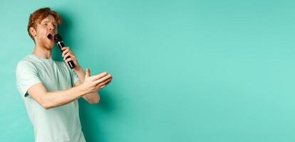Passionate redhead man in t-shirt singing serenade with microphone, looking aside at karaoke and gesturing, standing over mint background photo