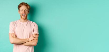 Enthusiastic redhead man in t-shirt checking out interesting promo, cross arms on chest and looking at camera with awe, standing over turquoise background photo