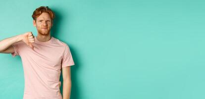 Disappointed redhead man showing thumbs-down, frowning and looking skeptical, epxress dislike and disapproval, standing over turquoise background photo