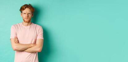 Young man with ginger hair and beard looking offended, feeling insulted and staring at camera, sulking at you, cross arms on chest defensive, standing over turquoise background photo