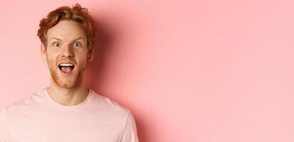 Headshot of amazed redhead male model in t-shirt open mouth, looking fascinated and happy at camera, standing over pink background photo